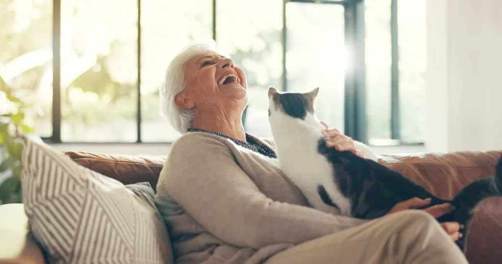 A resident enjoys a moment of laughter and pet interaction on a couch in a bright community setting.